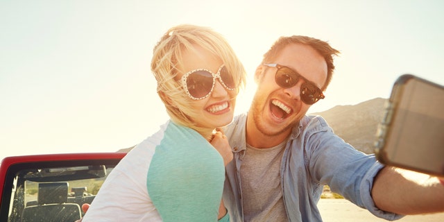 Couple On Road Trip Sit On Convertible Car Taking Selfie
